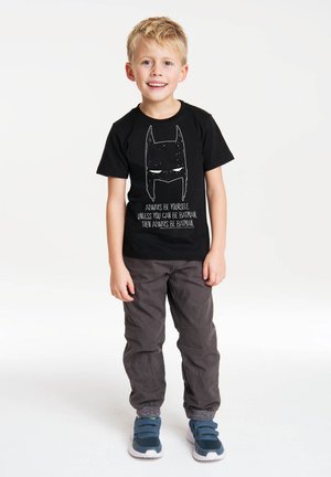 Schwarzes T-Shirt mit einer weißen Grafik eines Superheldengesichts und Text. Graue Hosen mit elastischen Bündchen. Blaue Sneakers mit Klettverschlüssen.