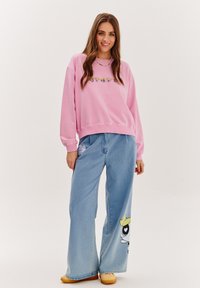 Růžová mikina s vyšívaným květinovým detailem, spárovaná s širokými světle modrými džíny s grafikou kreslené postavičky. Viditelné oranžové obuv.