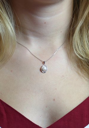 Zilveren ketting met een ronde parel pendant omringd door een gedetailleerd halo-ontwerp, liggend op blote huid tegen een rode stoffen achtergrond.