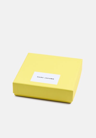 Kollane ristkülikukujuline kingitus karp, millel on sile pind, ning valge silt, kus on mustades paksudes tähtedes kirjas "MARC JACOBS".