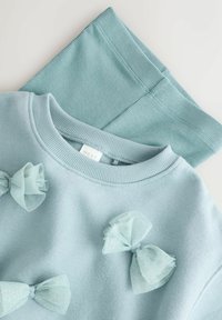 Lyseblå sweatshirt med sløjfer i gennemsigtigt materiale foran, parret med matchende ribbede leggings i et blødt stof.