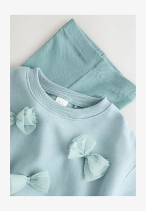 Gaiši zilais džemperis ar caurspīdīgā materiāla lentītēm priekšpusē, saskaņots ar atbilstošām ribotām legingām no mīksta auduma.