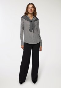 Camicia a righe bianco e nera con un maglione grigio annodato al collo, abbinata a pantaloni neri a gamba larga e scarpe nere.