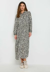 Schwarz-weißblumengemustertes langes Kleid mit Kragen, langen Ärmeln, plissierter Gestaltung und getragen mit hellen Sneakers.
