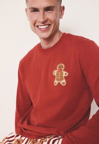 Rode longsleeve met een gingerbread man-applicatie in goud en bruin. Gemaakt van katoen met een ronde hals en een losse pasvorm.