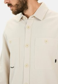 Beige skjorte med knapper lavet af tekstureret stof, med to brystlommer, beige knapper og en sort detalje nær venstre lomme.