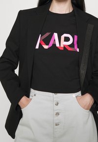 Persona vestida con una blazer negra sobre una camiseta corta negra con el texto colorido "KARL", metida en unos pantalones gris claro de cintura alta con botones.