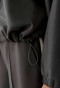Schwarze Sportjacke mit gerafftem Bündchen und Saum, ausgestattet mit einem verstellbaren Kordelzug mit metallic Toggle. Glatter, leicht glänzender Stoff.