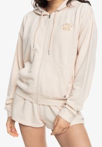 Donna che indossa una felpa con zip beige chiaro e pantaloni corti abbinati, con il logo "ROXY" e ricami floreali sul petto.