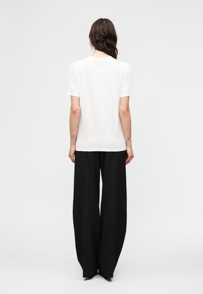 Γυναίκα με μακριά σκούρα μαλλιά που φορά ένα απλό λευκό κοντομάνικο μπλουζάκι και μαύρο παντελόνι φαρδύ, στέκεται γυρισμένη με την πλάτη σε λευκό φόντο.