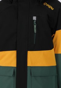 Veste noire, jaune et verte avec une poche zippée sur la poitrine, deux poches à rabat en bas, et un logo ZigZag jaune sur l'épaule.