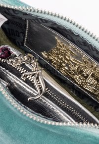 Inre delen av en teal mockapåse som visar ett silverfärgat dragkedjefack, gyllene utsmyckningar och en silverkedja med en dekorativ berlock.