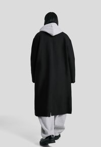 Zwarte oversized jas met een grijze hoodie eronder. Heeft brede mouwen en een rechte rug. Grijze joggingsbroek en zwarte schoenen maken de look compleet.