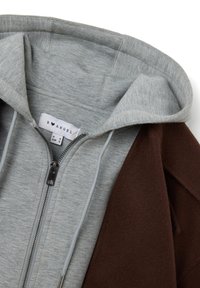 Sudadera con cremallera gris con un interior suave, que presenta una capa exterior marrón. Incluye una capucha con cordón y una etiqueta de marca en el interior.