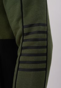 Λεπτομέρεια μανικιού πράσινου υφάσματος με επτά οριζόντιες μαύρες ρίγες και μαύρο περιθώριο κατά μήκος της ραφής.