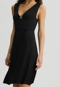 Musta hihaton mekko, jossa V-kaula-aukko, kultaiset yksityiskohdat rinnalla ja kiristysnauha vyötäröllä, laskeutuu kevyesti lanteilta. Pehmeä kangaspinta.