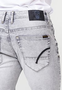 Grå denim jeans med et unikt design og mørke stikninger. Baglommerne har en særlig form, og et mærket lædermærke er synligt.