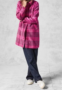 Abrigo de lana a cuadros rosas con cuello, cierre de botones y cinturón; combinado con vaqueros oscuros y zapatillas blancas. Textura suave y cálida.