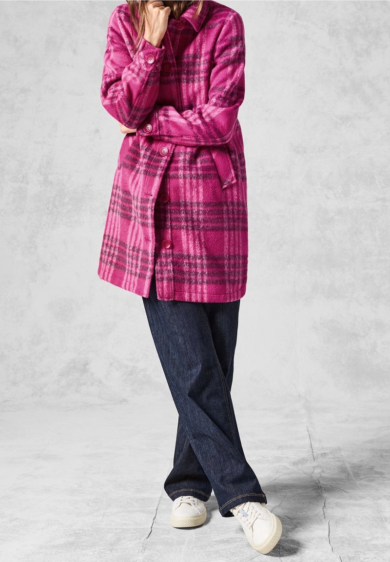 Abrigo de lana a cuadros rosas con cuello, cierre de botones y cinturón; combinado con vaqueros oscuros y zapatillas blancas. Textura suave y cálida.