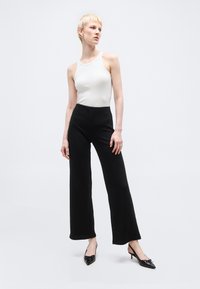 Άτομο που φοράει λευκό αμάνικο τοπ, μαύρο παντελόνι φαρδύ στα πόδια και μαύρες γόβες με ακτίνες και λουράκι πίσω, στέκεται μπροστά σε απλό ανοιχτό φόντο.