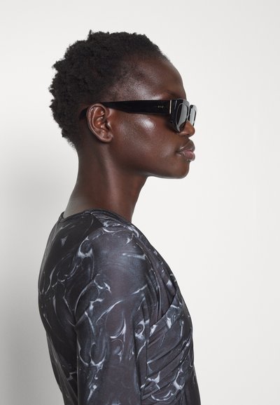 Čierne slnečné okuliare s hrubým rámom, nosené osobou v sivom mramorovom topu. Vlasy sú krátke a textúrované. Profilový pohľad na bielom pozadí.