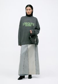 Suéter cinza oversized com o texto verde "STIEGLITZ EST.2014". Combinado com uma saia de ganga longa em bloco de cor e botas de salto pretas. Bolsa preta com tachas.