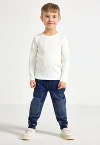 Long-sleeve white ribbed shirt with blue denim pants featuring elastic cuffs, classic five-pocket design, and beige sneakers.