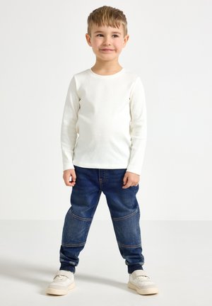 Wit ribbed shirt met lange mouwen, gecombineerd met blauwe denim broek met elastische manchetten, klassieke vijf-pocket ontwerp en beige sneakers.