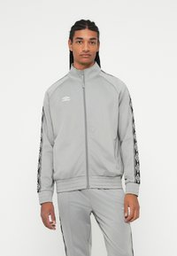 Hombre con cabello trenzado que lleva un chándal de Umbro de color gris claro con rayas de patrón negro y blanco en las mangas y piernas del pantalón, de pie mirando al frente.