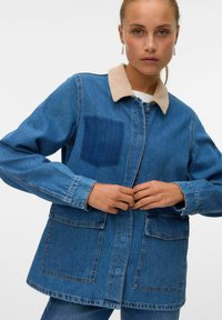 Denim jacket in blue with a beige corduroy collar, featuring a patch pocket, button closure, and two front pockets. Collar is textured.