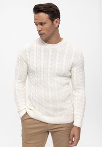 Suéter blanco de punto con patrón de ochos, cuello redondo y puños y dobladillo acanalados. Combinado con pantalones beige. Textura suave, diseño ajustado.