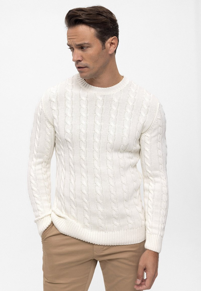 Suéter blanco de punto con patrón de ochos, cuello redondo y puños y dobladillo acanalados. Combinado con pantalones beige. Textura suave, diseño ajustado.