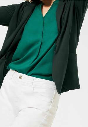 Person, die eine grüne Bluse, einen schwarzen Blazer und weiße Hose trägt, von der Mitte des Oberkörpers aufwärts vor einem einfarbigen Hintergrund gezeigt.