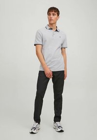 Hellgraues Poloshirt mit einem schwarzen Kragen, kombiniert mit dunkler Hose. Das Modell steht in neutraler Pose und trägt weiße und schwarze Turnschuhe.