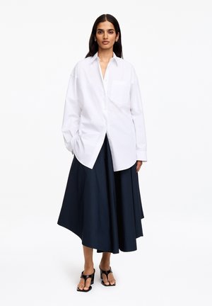 Femme portant une chemise blanche ample boutonnée, une jupe midi bleu marine et des sandales noires à talons, debout devant un fond blanc uni.