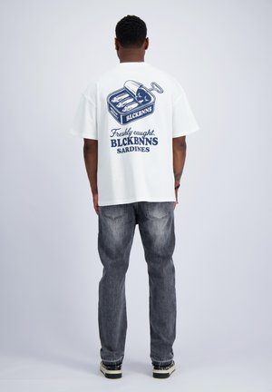 Mann steht mit dem Rücken zur Kamera, trägt ein weißes T-Shirt mit einer Sardinendosen-Grafik und dem Text "Freshly caught BLCKBNNS SARDINES" sowie ausgewaschene Jeans.