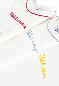 Tre vita bomullskläder med olika färgade broderade fraser: röd "Petit amour", blå "Petit ange" och gul "Petit cœur."