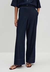 Wide-leg dark denim trousers with side pockets, contrasting stitching, and a smooth texture. Paired with flip-flops on a neutral background.