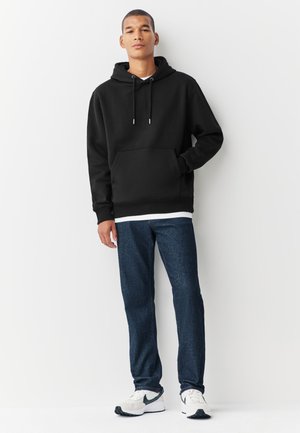Ung mand iført sort hættetrøje, blå jeans og hvide sneakers med sorte accenter, står mod en hvid baggrund.