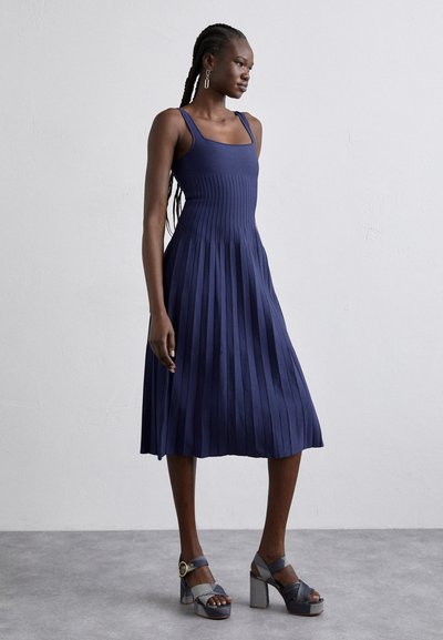 Marineblaues plissiertes Kleid mit quadratischem Ausschnitt und Spaghetti-Trägern. Kombiniert mit mehrfarbigen Plateausandalen mit einem dicken Absatz.