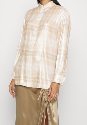 Femme portant une chemise à carreaux beige brillante avec boutons et une jupe beige soyeuse avec une fente latérale froncée, debout sur un fond blanc.