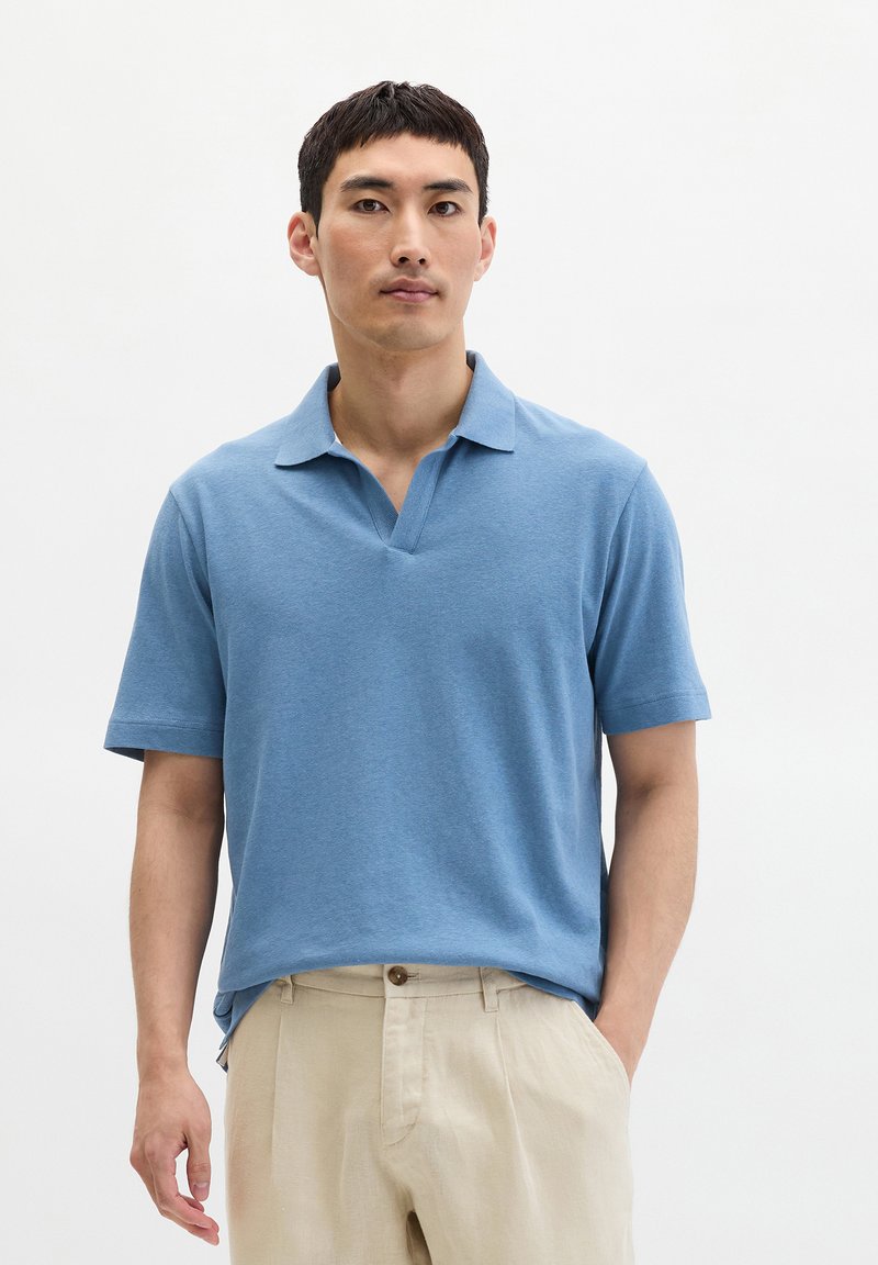 Mann trägt ein blaues kurzärmeliges Poloshirt und beige Hose, steht mit einer Hand in der Tasche vor einem einfarbigen weißen Hintergrund.