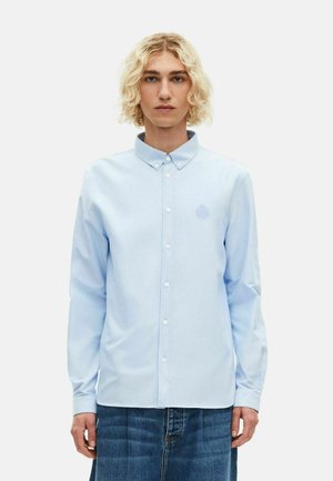 Camisa de manga larga azul claro con cuello de botón, presenta un sutil logo bordado en el pecho y una textura suave de algodón.