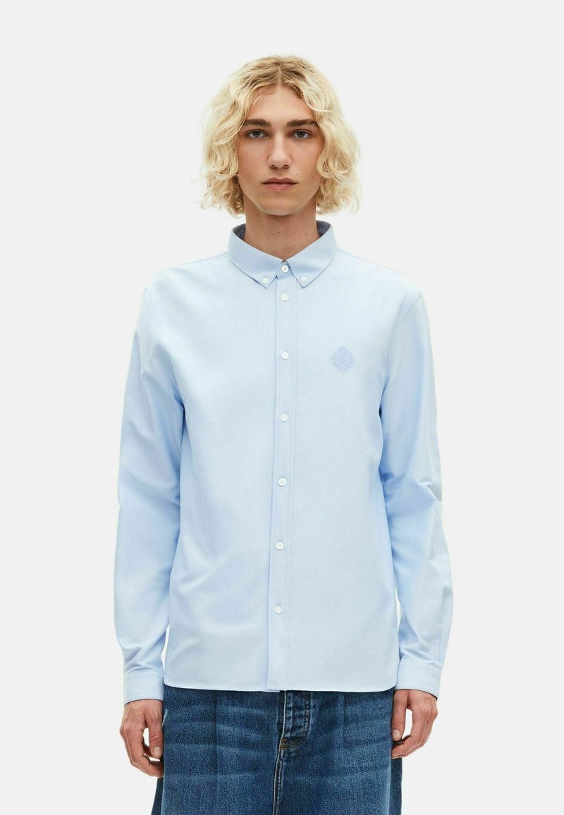 Camisa de manga larga azul claro con cuello de botón, presenta un sutil logo bordado en el pecho y una textura suave de algodón.