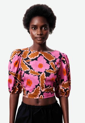 Femme portant un chemisier court à motif floral rose, orange et marron avec des manches bouffantes jusqu'aux coudes, assorti à un pantalon noir.