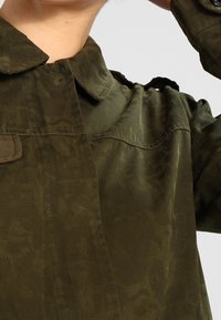 Giacca verde oliva con una texture liscia, dotata di colletto, tasca frontale e dettagli sulle spalle, realizzata in un motivo sottile.