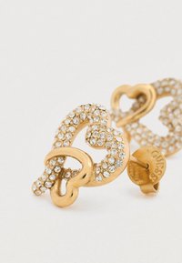 Σκουλαρίκια σε χρυσαφί τόνο, σχηματισμένα σαν διαπλεκόμενες καρδιές, διακοσμημένα με καθαρά κρύσταλλα και με ασφαλή πίσω μέρος με λεπτομέρειες σήμανσης.