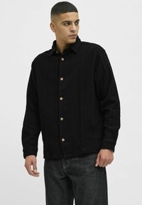 Chaqueta negra de botones con una tela texturizada, con un collar y botones de madera. Combinada con jeans oscuros, mostrando un ajuste relajado.