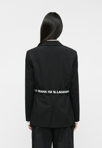 Μαύρο σακάκι από απαλό ύφασμα, με προσαρμοσμένη εφαρμογή, κέντρα lapels και ελαστική ζώνη με σήμα στη μέση με λευκό κείμενο.