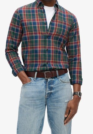 Mann trägt ein grün-rotes-blau kariertes Hemd über einem weißen T-Shirt, hellblaue Jeans, braunen Ledergürtel und eine schwarze Armbanduhr.